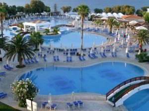 Basen z wieloma parasolami i leżakami, otoczony palmami i budynkami. Traveligo Blog