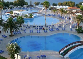 Basen z wieloma parasolami i leżakami, otoczony palmami i budynkami. Traveligo Blog