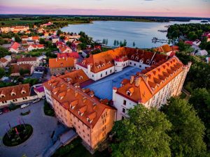 Zdjęcie przedstawia widok zamku Ryn z lotu ptaka, położonego nad jeziorem w miasteczku Ryn. Zamek z czerwonymi dachami, otoczony drzewami, znajduje się na tle malowniczego zachodu słońca. - Traveligo Blog