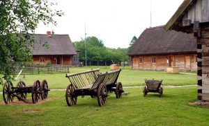 Drewniane domy z dachem ze strzechy. Stare kolorowe wozy zaprzęgowe. Trawnik. Traveligo Blog
