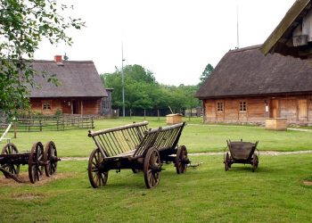 Drewniane domy z dachem ze strzechy. Stare kolorowe wozy zaprzęgowe. Trawnik. Traveligo Blog