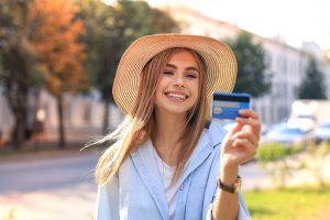 Kobieta uśmiechnięta, trzymająca kartę kredytową, ubrana w niebieską kurtkę i kapelusz. Traveligo Blog