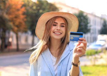 Kobieta uśmiechnięta, trzymająca kartę kredytową, ubrana w niebieską kurtkę i kapelusz. Traveligo Blog
