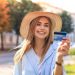 Kobieta uśmiechnięta, trzymająca kartę kredytową, ubrana w niebieską kurtkę i kapelusz. Traveligo Blog