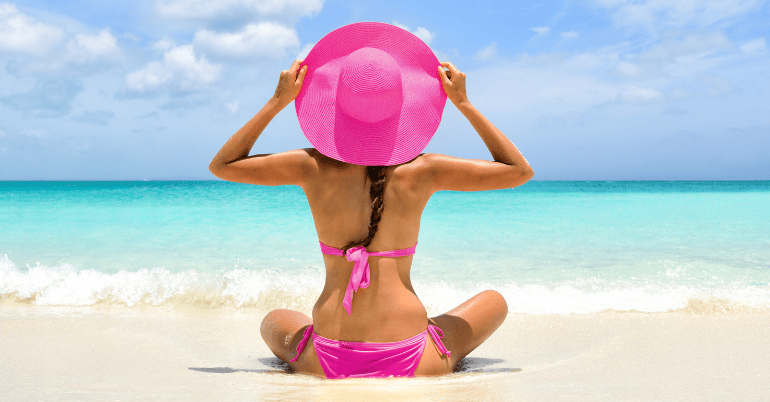 kobieta w różowym bikini i kapeluszu siedzi na plaży z widokiem na turkusowe morze
